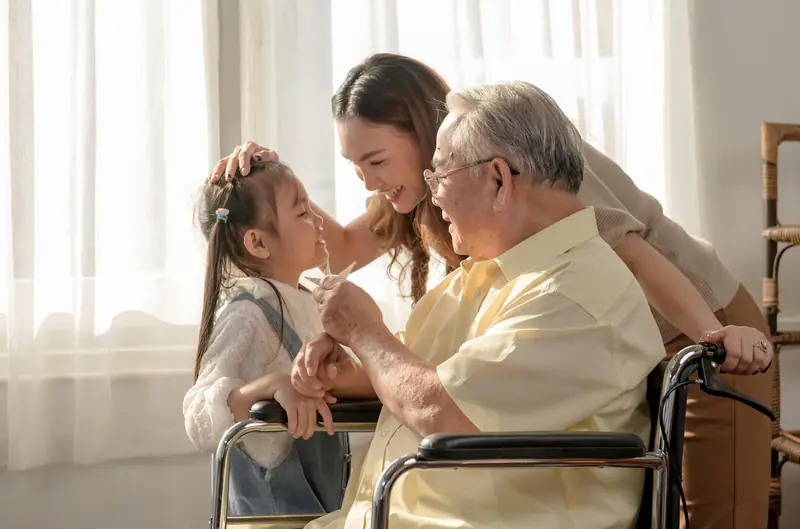 A grandfather in a wheelchair sharing a tender moment with his daughter and granddaughter at home.