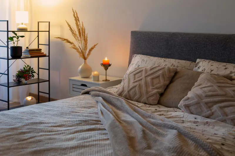 A warm, softly lit bedroom at home with a freshly made bed and a candle on the nightstand.