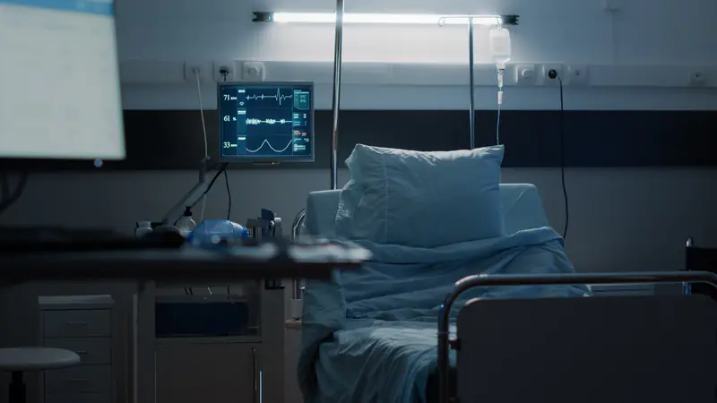 An empty hospital bed beside a bedside monitor in a dimly lit ward.
